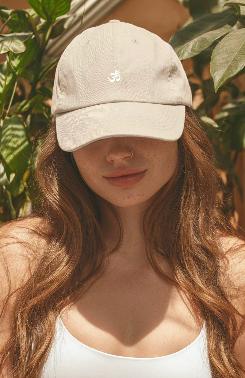 Spiritual woman wearing a beige baseball cap with an Om logo, standing among green plants.